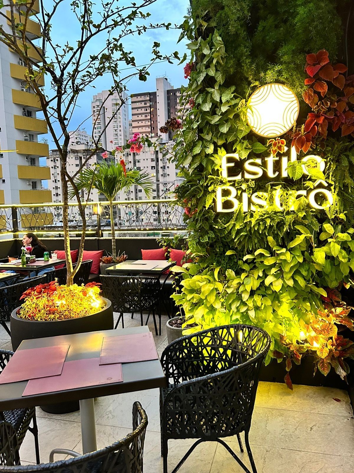 Rooftop do Estilo Bistrô com o letreiro entre o jardim vertical, mesas e vista da cidade